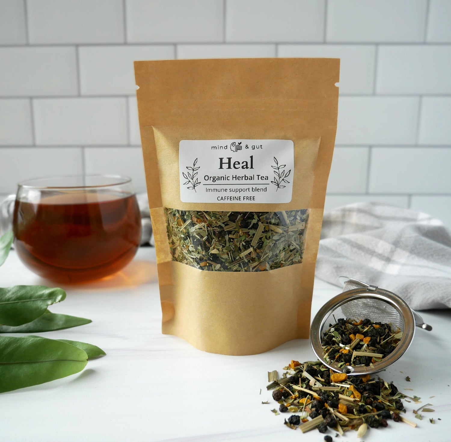 Bag of 'Heal' organic herbal tea with a glass of tea and loose tea leaves on a white surface.