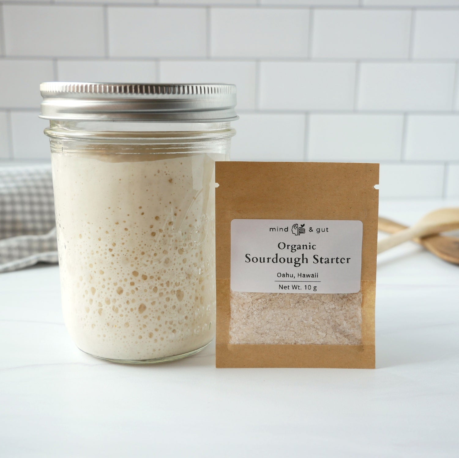 Jar of active organic sourdough starter next to dehydrated organic starter for sale on a marble kitchen counter. 