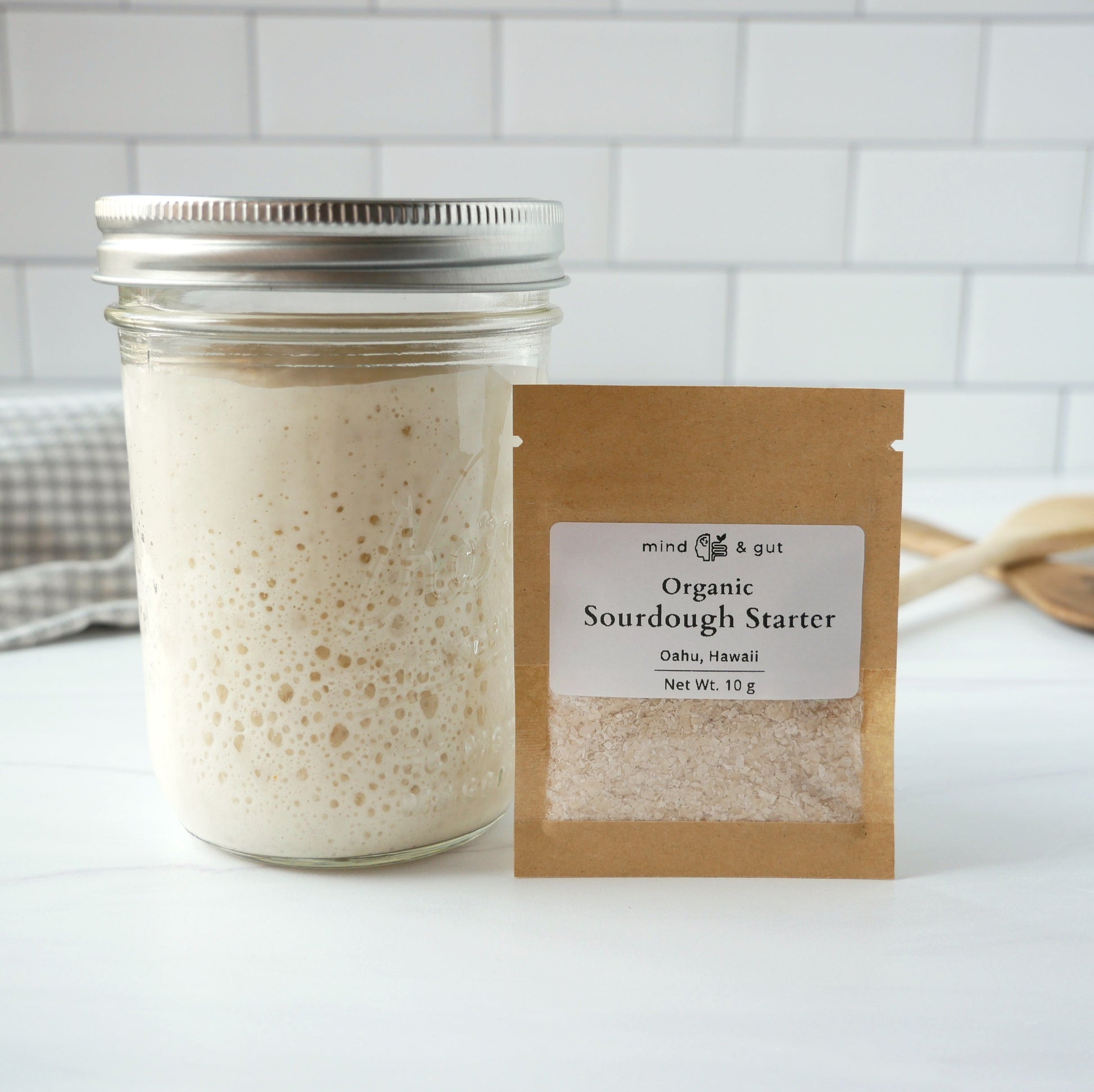 Jar of active organic sourdough starter next to dehydrated organic starter for sale on a marble kitchen counter. 